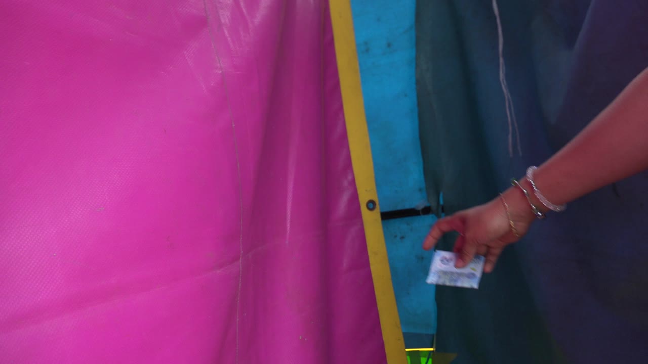Close frame in San Jose del Cabo showing a pink plastic tarp with yellow edging, a blue support panel, dark fabric, and a bracelet-wearing hand holding a small paper at the right, with no visible faces.