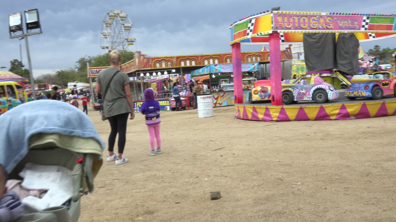 Woman and child facing colorful amusement rides on a dirt fairground.