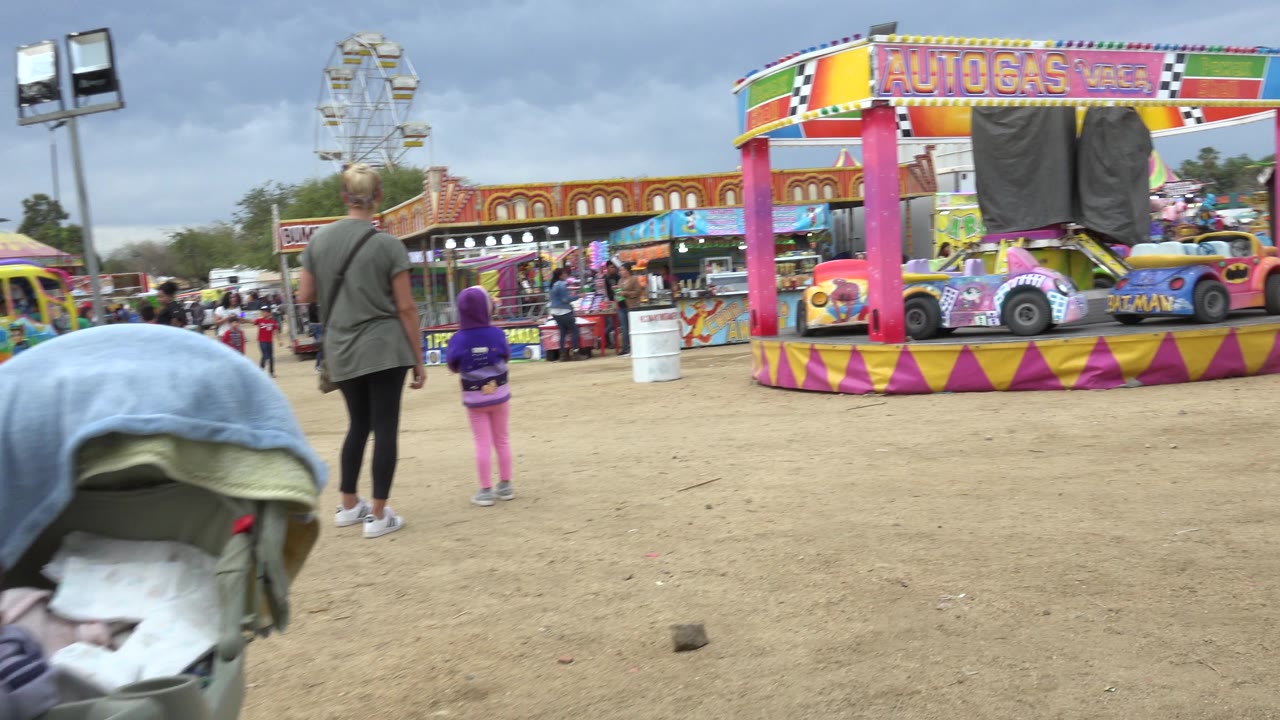 The woman and child stand farther into the fairground with a colorful Autogas ride canopy, bumper cars, a Ferris wheel, and visitors visible in San Jose Del Cabo, Mexico.