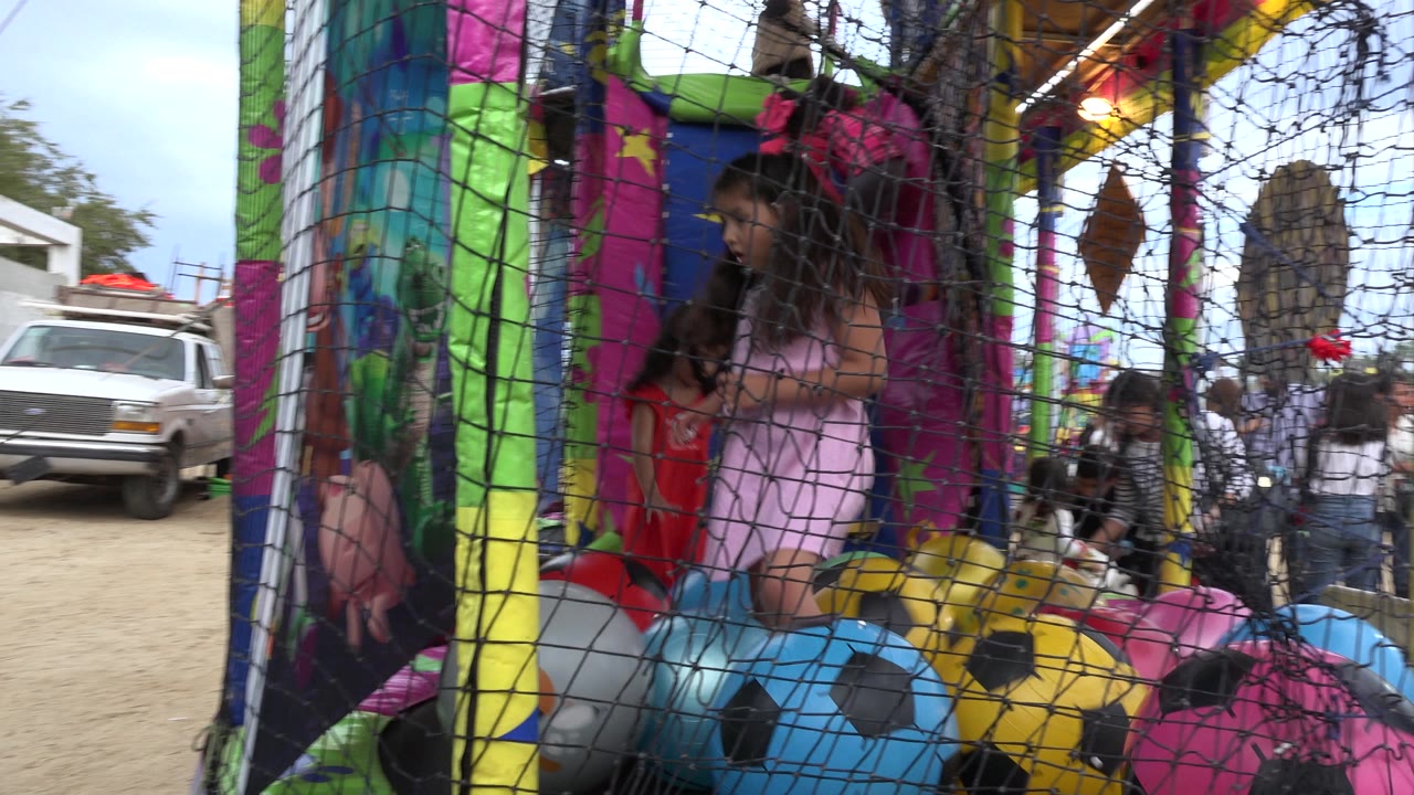 A girl in a pink dress stands behind black trampoline netting among colorful soccer-pattern balls at a fair attraction in San Jose Del Cabo, Mexico.