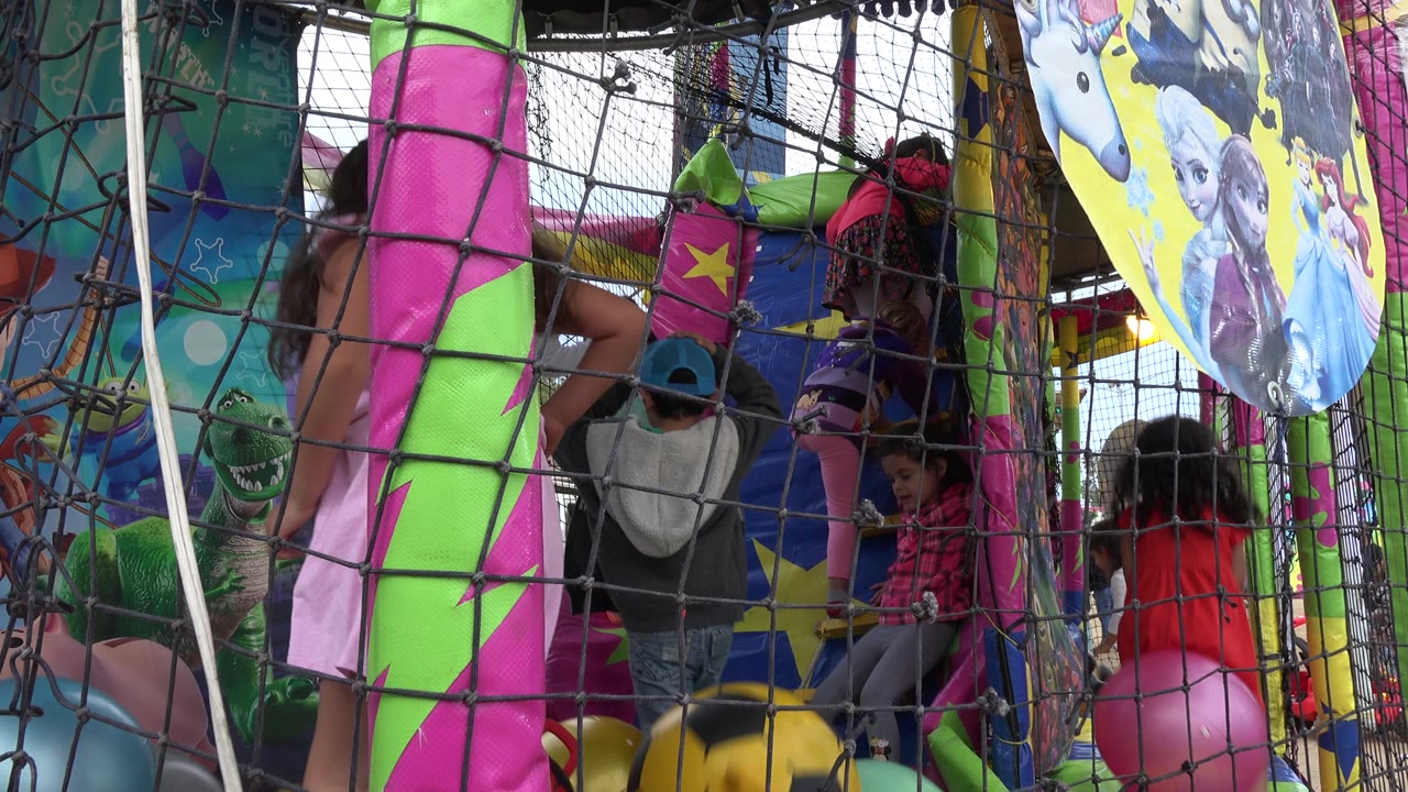 Children play inside a colorful inflatable playground in San Jose del Cabo, Mexico, with black netting in the foreground and cartoon panels above the bounce area.