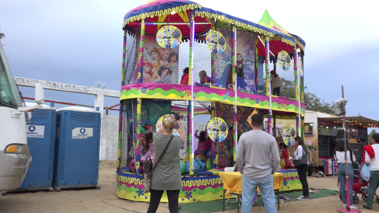 At 0:00 in San Jose del Cabo, children are visible inside a bright two-level oval play structure while adults stand in front and a white bus and blue portable toilets sit behind it.