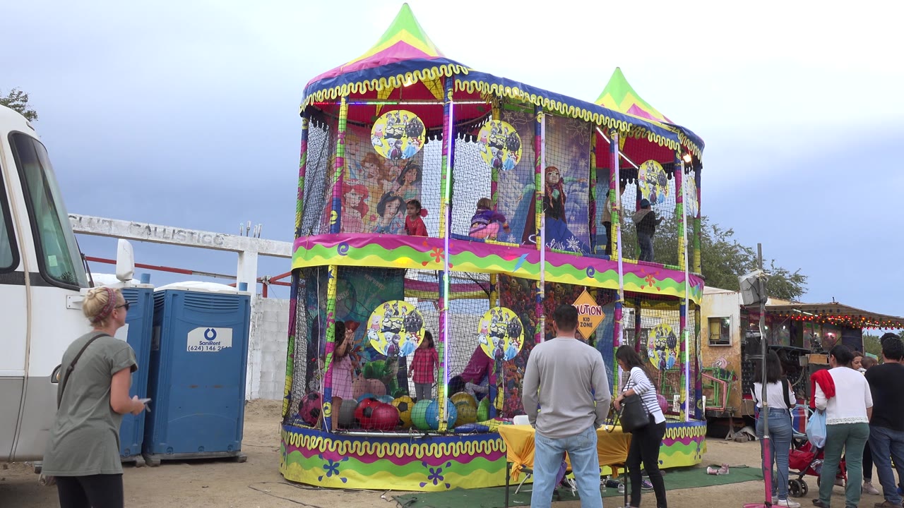 The San Jose Del Cabo amusement attraction fills the frame, with children behind mesh panels, adults in the foreground, and a white bus and portable toilets behind the structure.