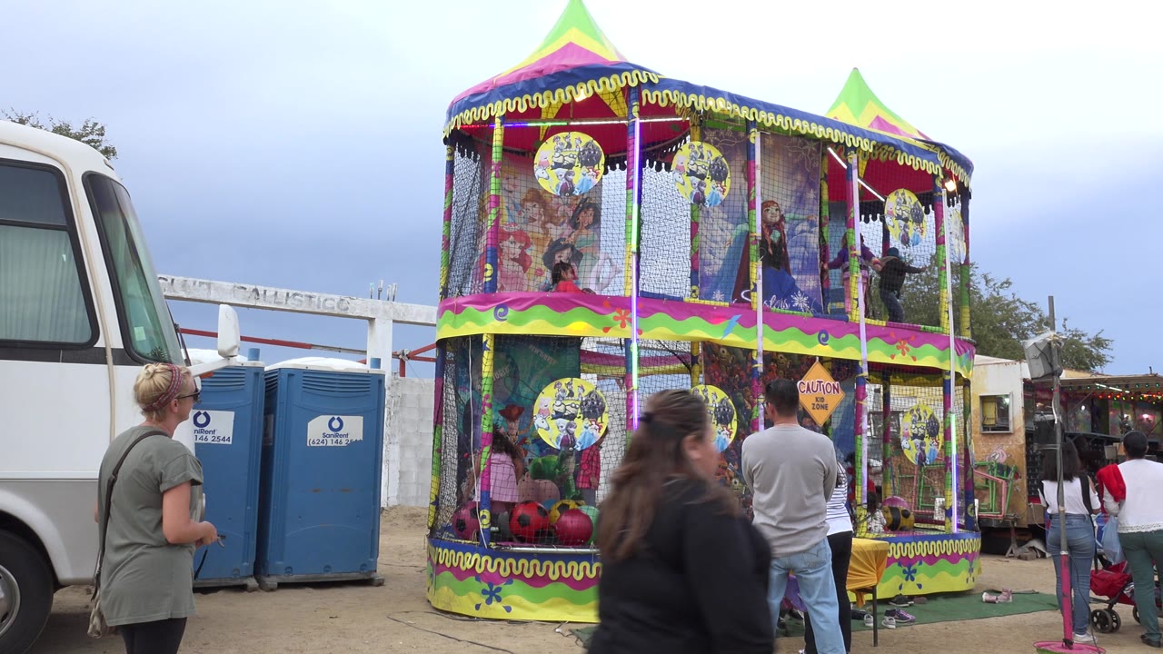 The San Jose del Cabo bounce house remains centered, with a man facing the inflatable, a woman near the portable toilets, children visible on both levels, and a passerby crossing the foreground.