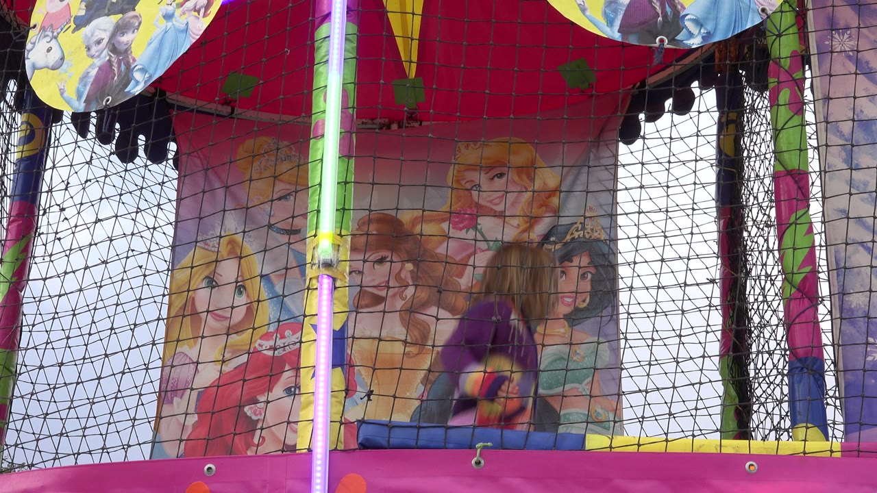 A child in a purple top is mid-bounce behind black trampoline netting in San Jose Del Cabo, Mexico, with pink trim and cartoon princess artwork behind them.