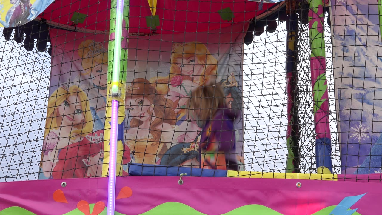 At 0:01 in San Jose del Cabo, Mexico, the girl is motion-blurred beside a bright vertical trampoline pole, with safety netting crossing the frame and character artwork behind her.