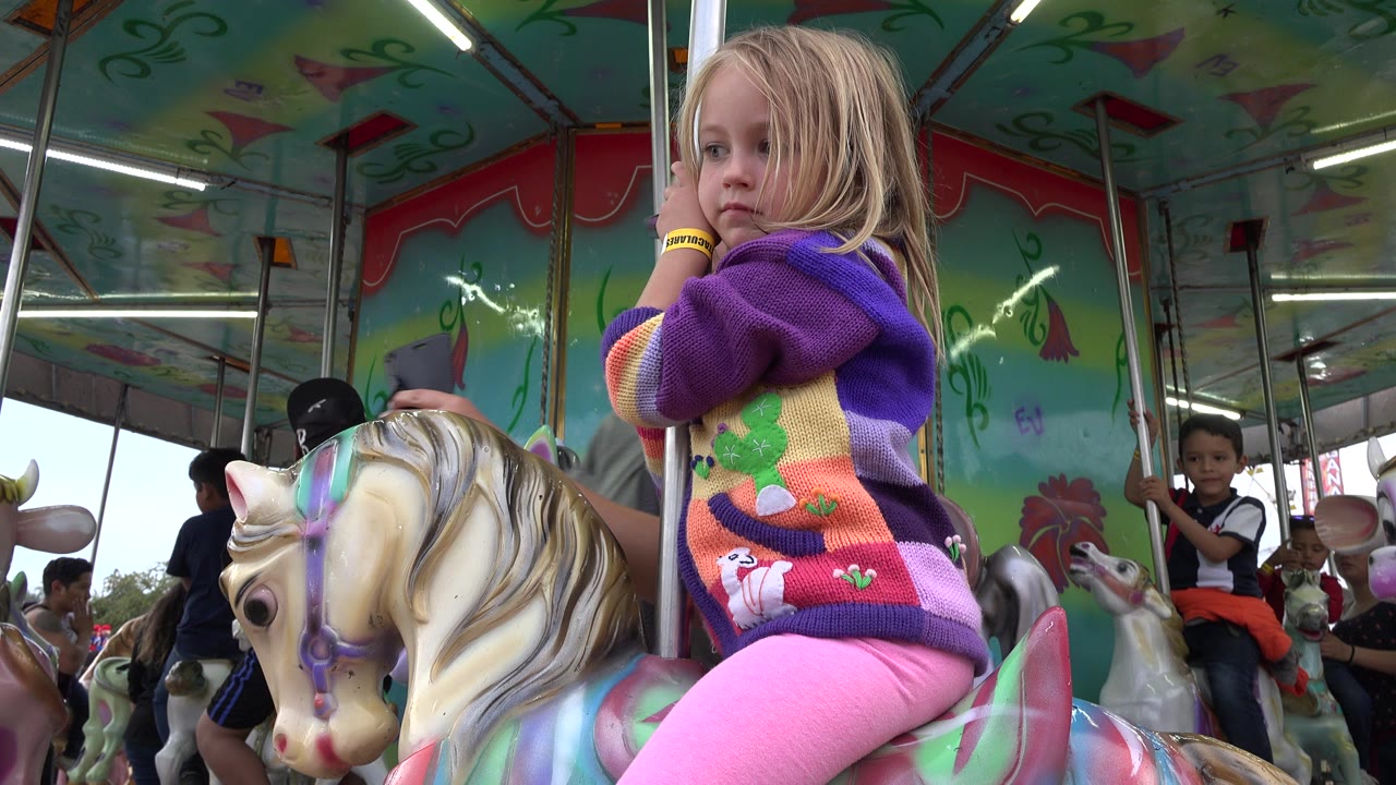 A blonde child in a purple sweater sits on a painted mechanical horse at a fair ride in San Jose del Cabo, Mexico, with other riders partly visible behind.