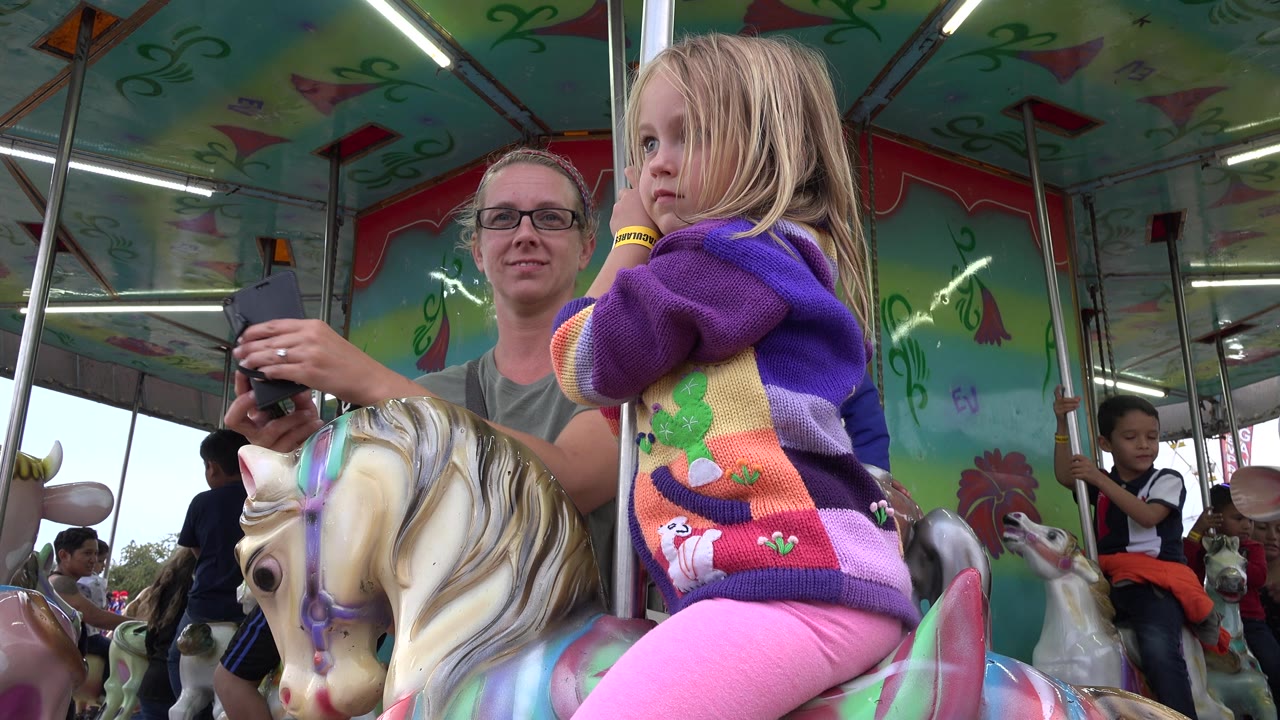Adult beside child on mechanical horse holds a phone near the ride pole.