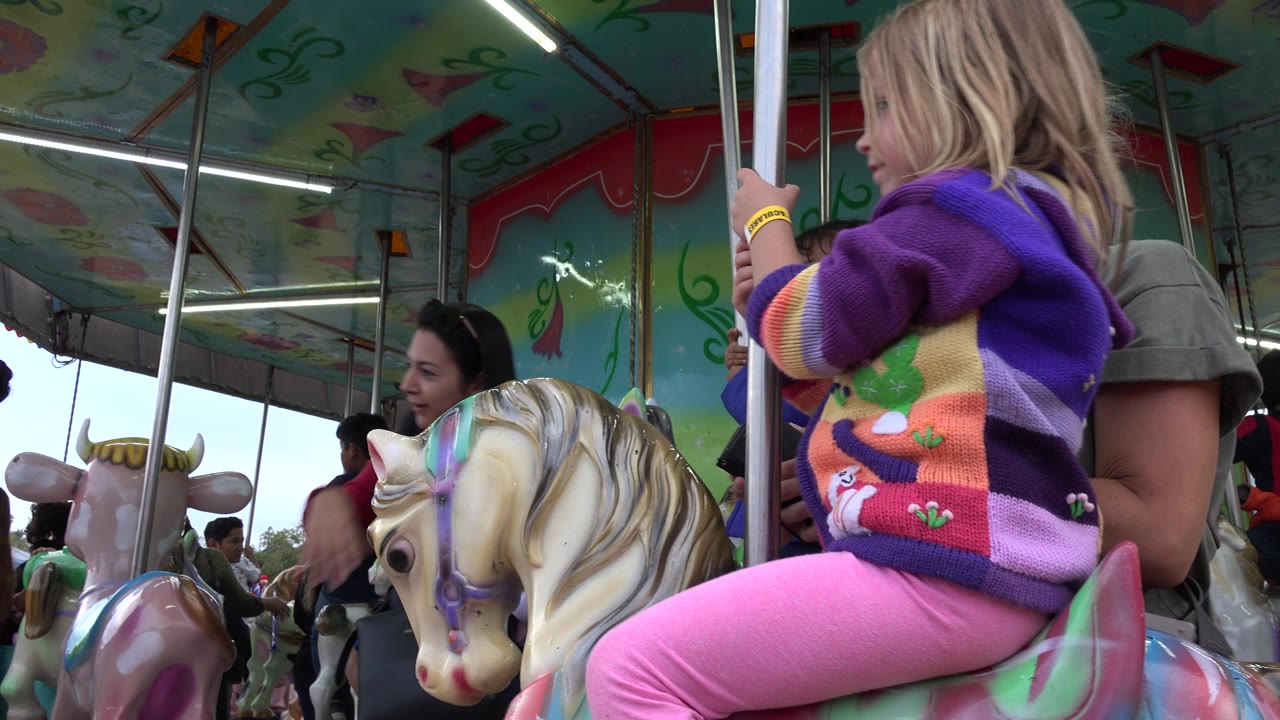 A child in pink pants and a purple striped sweater sits on a carousel horse in San Jose del Cabo, Mexico, with adults and other riders nearby under a colorful canopy.