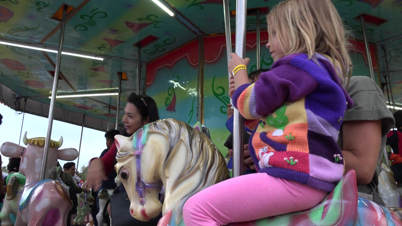 The child remains seated on the painted carousel horse in San Jose del Cabo, Mexico, holding the pole as another child and adults appear in the background.