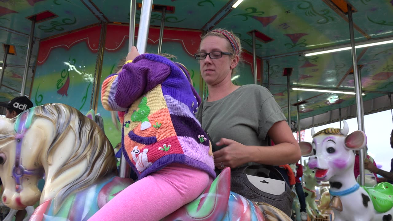 The child leans toward the carousel pole with the adult woman close behind, surrounded by painted horses and animal figures on the San Jose del Cabo ride.