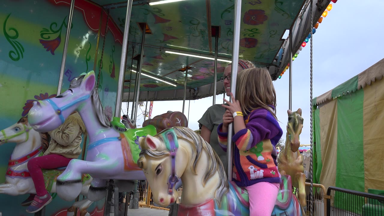 Mother and daughter beside painted carousel horses under a green canopy.