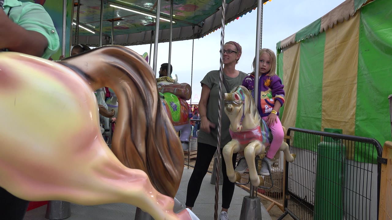 In San Jose del Cabo, Mexico, a child sits on a small painted amusement-ride animal beside an adult while a large blurred carousel horse fills the left foreground.