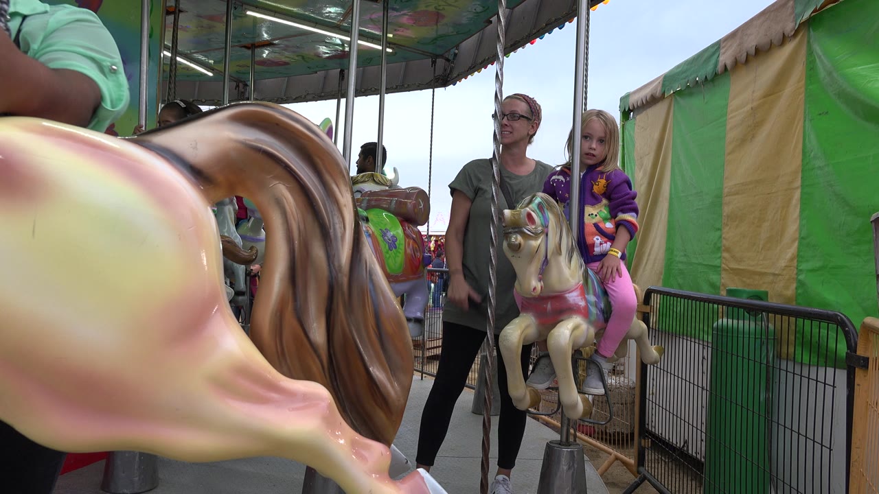 The child remains seated on the small ride figure in San Jose del Cabo, Mexico, with the adult standing close by and a blurred carousel horse still crossing the foreground.
