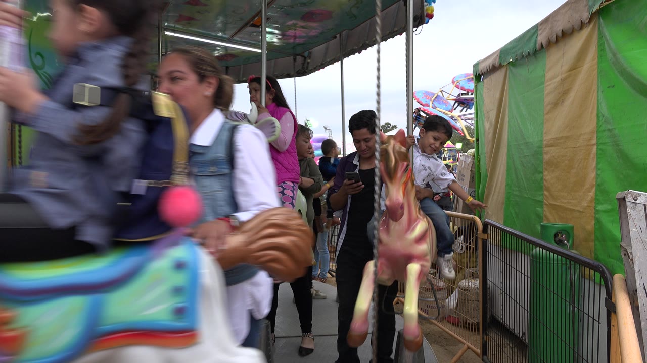 At the start of the San Jose Del Cabo clip, a blurred carousel rider and painted horse pass close to the camera while adults and children stand around the ride.