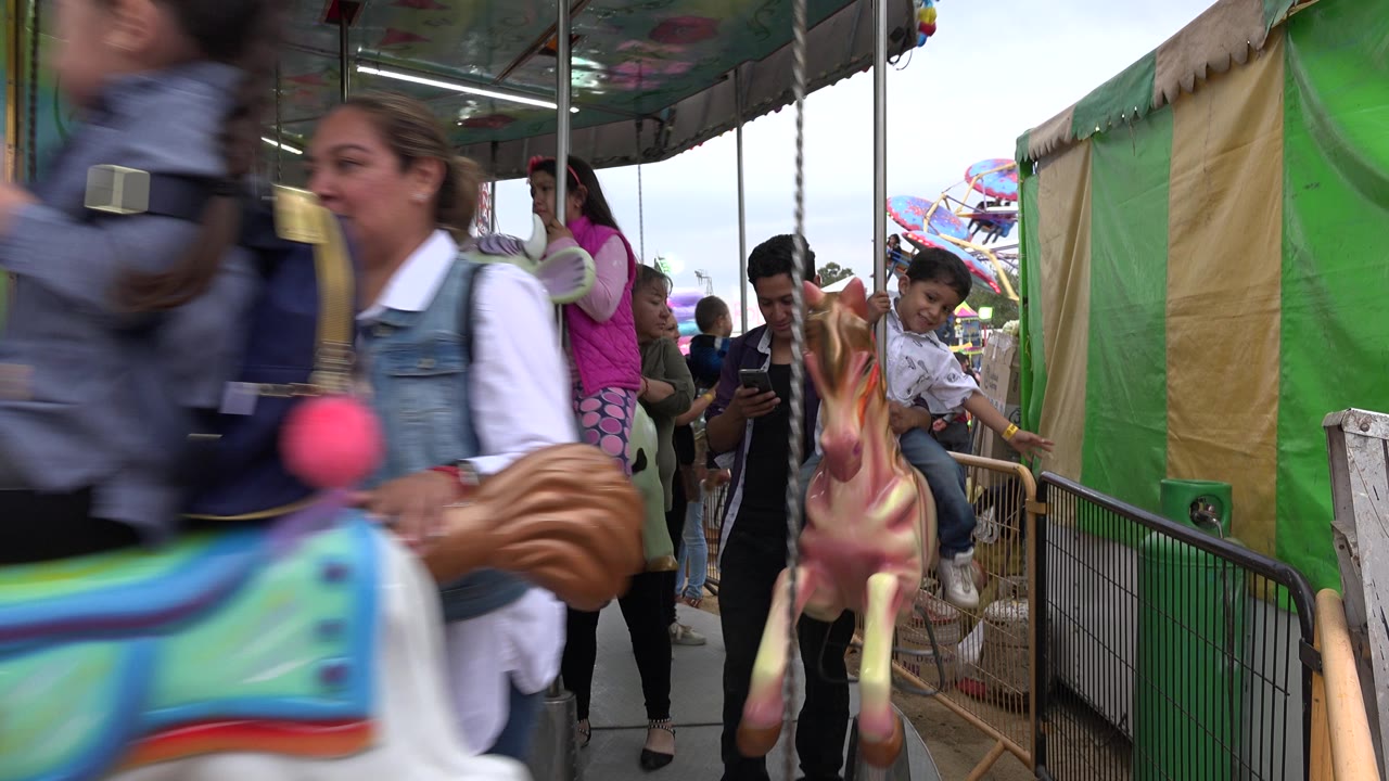 A child rides a painted carousel horse in San Jose Del Cabo while adults stand inside and beside the ride under green and tan canvas panels.