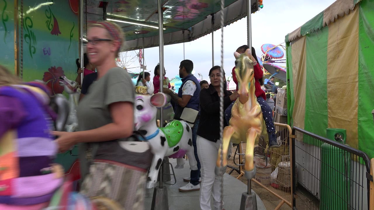 A woman walks past the foreground of a carousel in San Jose Del Cabo, Mexico, while painted animal figures and waiting visitors fill the ride platform.