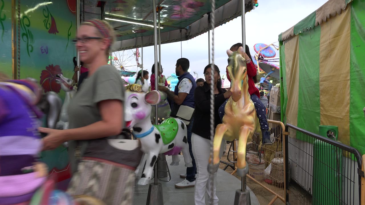 Carousel animals pass the camera beside green painted panels in San Jose Del Cabo, Mexico, with adults and children visible around the platform.