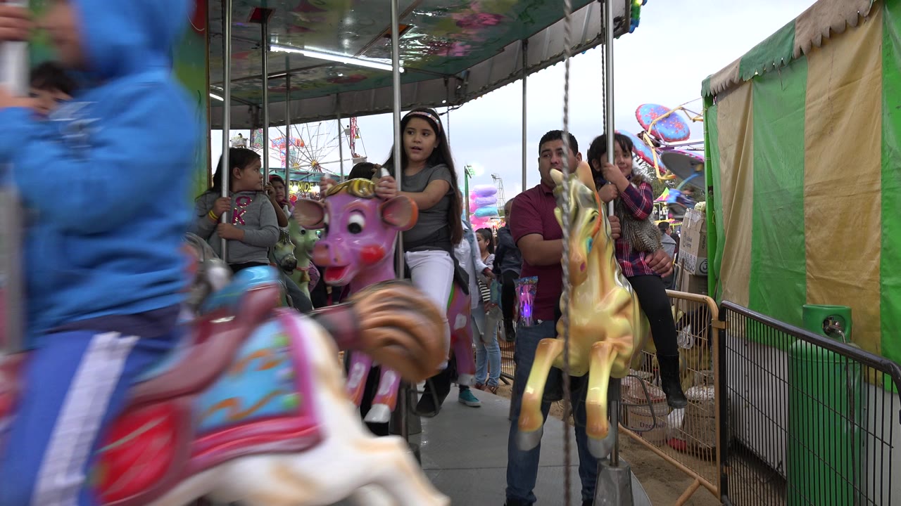 At the start of the clip in San Jose del Cabo, Mexico, children and adults ride a merry-go-round while a blue-clad rider and painted horse pass close to the camera.