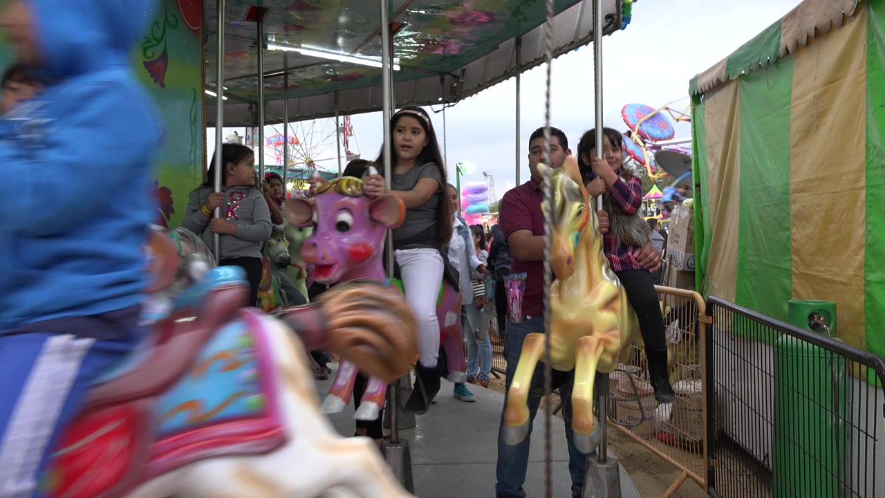 One second in, riders on painted carousel animals pass a fixed ground-level view in San Jose del Cabo, Mexico, with children and adults visible near the railing.