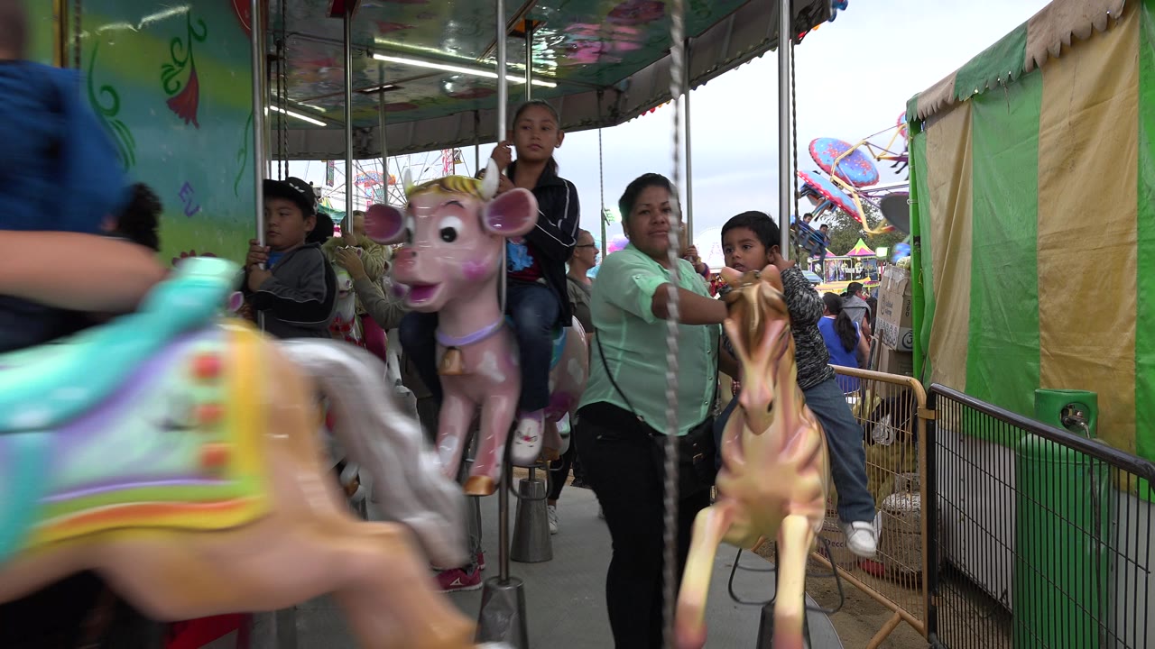 Children ride painted carousel horses in San Jose del Cabo, Mexico, while adults stand close by near the green ride panels and metal poles.