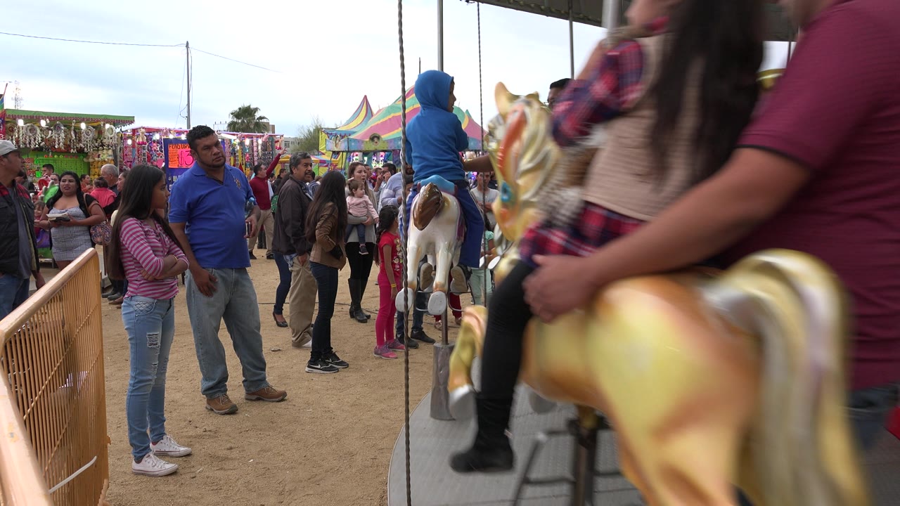A child riding a painted carousel horse passes close to the camera while families and fair booths remain visible behind the ride in San Jose Del Cabo, Mexico.