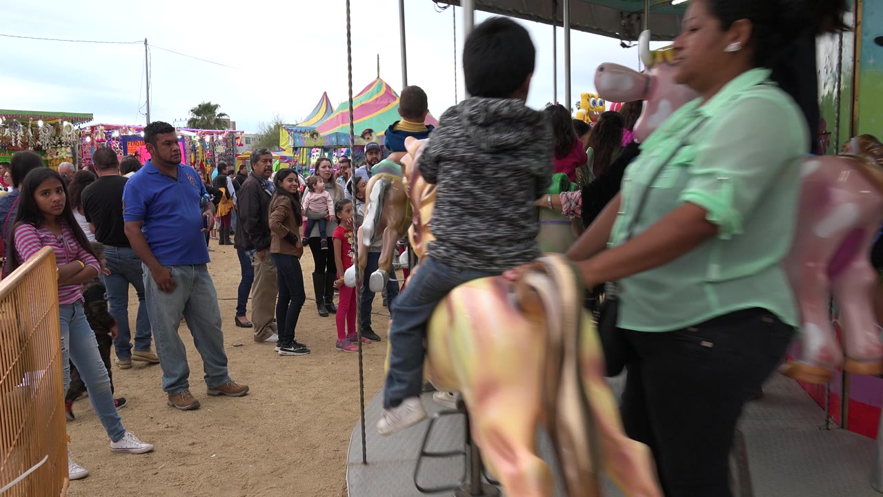 At 0:01, an adult in a light green shirt and a child on a carousel horse pass near the camera while fairgoers stand behind the railing in San Jose del Cabo, Mexico.