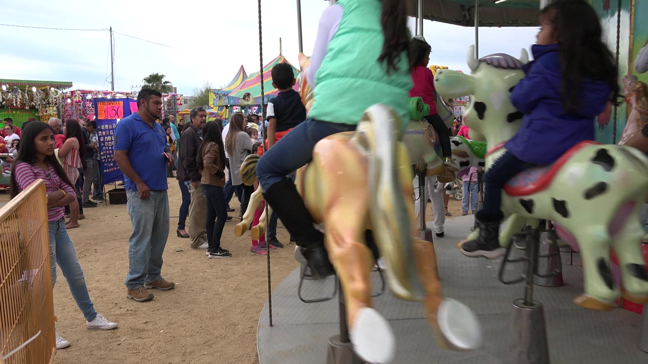 A carousel horse and child rider fill the foreground while people wait near booths and a colorful tent in San Jose del Cabo, Mexico.