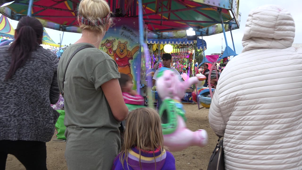 Adults and a child remain near the ride barrier in San Jose Del Cabo as a pink carousel animal passes through the center of the frame.