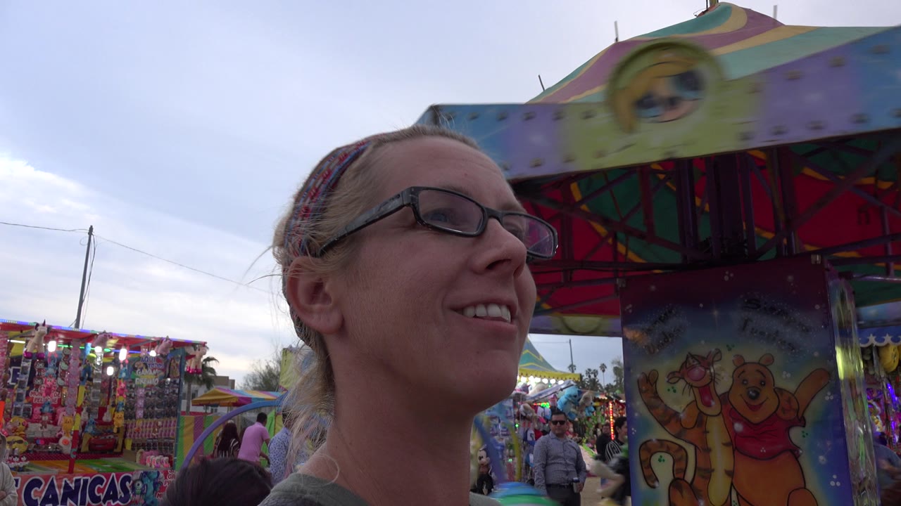 The woman remains close to camera as the ride's painted center column, cartoon characters, hanging prizes, and fairgoers fill the background in San Jose del Cabo, Mexico.