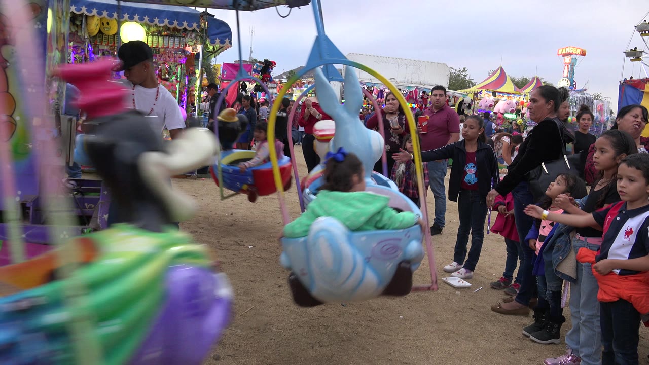 A San Jose Del Cabo carnival frame shows children riding animal-shaped swing seats past families waiting beside a purple ride structure.