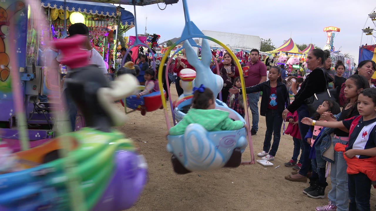 At one second, a bright carnival seat blurs across the foreground while adults and children stand near the San Jose Del Cabo ride platform.