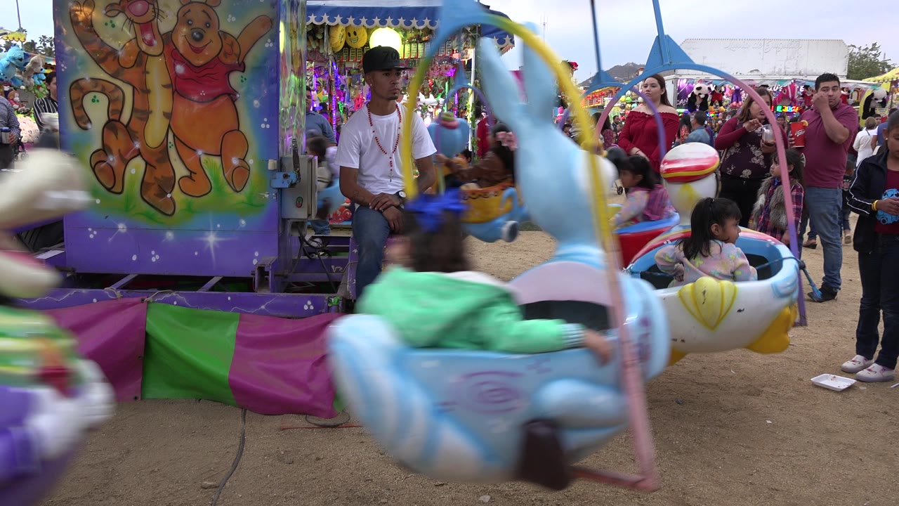 At 0:01, a blue carousel seat blurs across the foreground as the operator sits near the center and families watch from the crowded San Jose del Cabo fairground.
