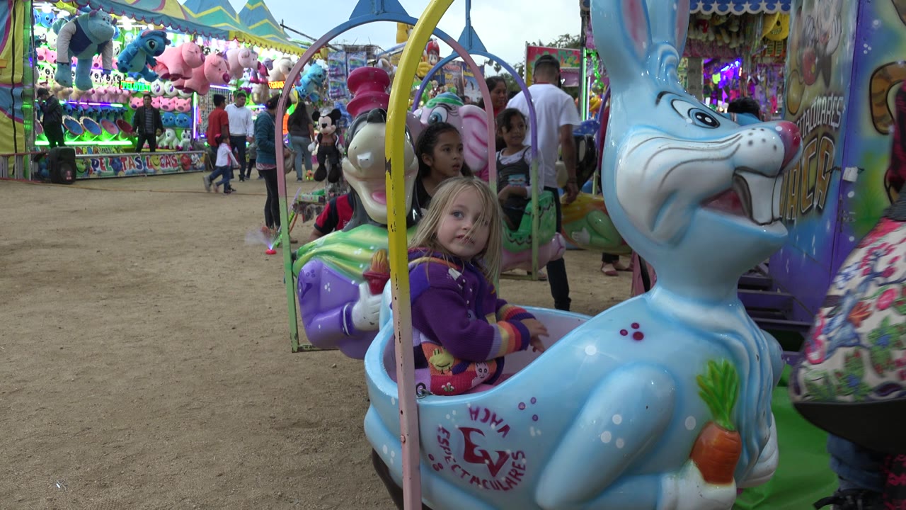 The blue rabbit ride car remains prominent as the child looks outward, with walking adults, other children, bright booth lights, and stuffed animal prizes visible behind her in San Jose del Cabo, Mexico.