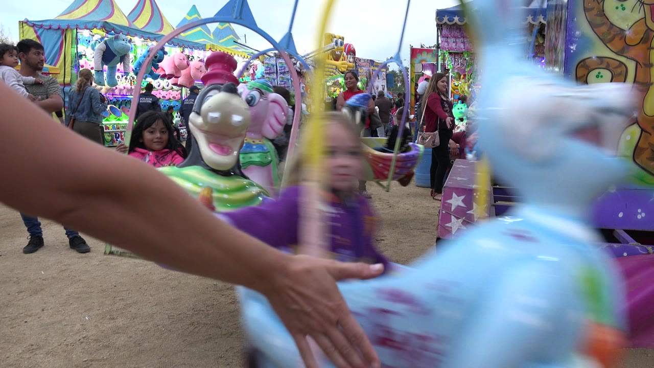 A blurred blue animal ride seat crosses the foreground as children and adults remain visible around the colorful ride in San Jose del Cabo, Mexico.