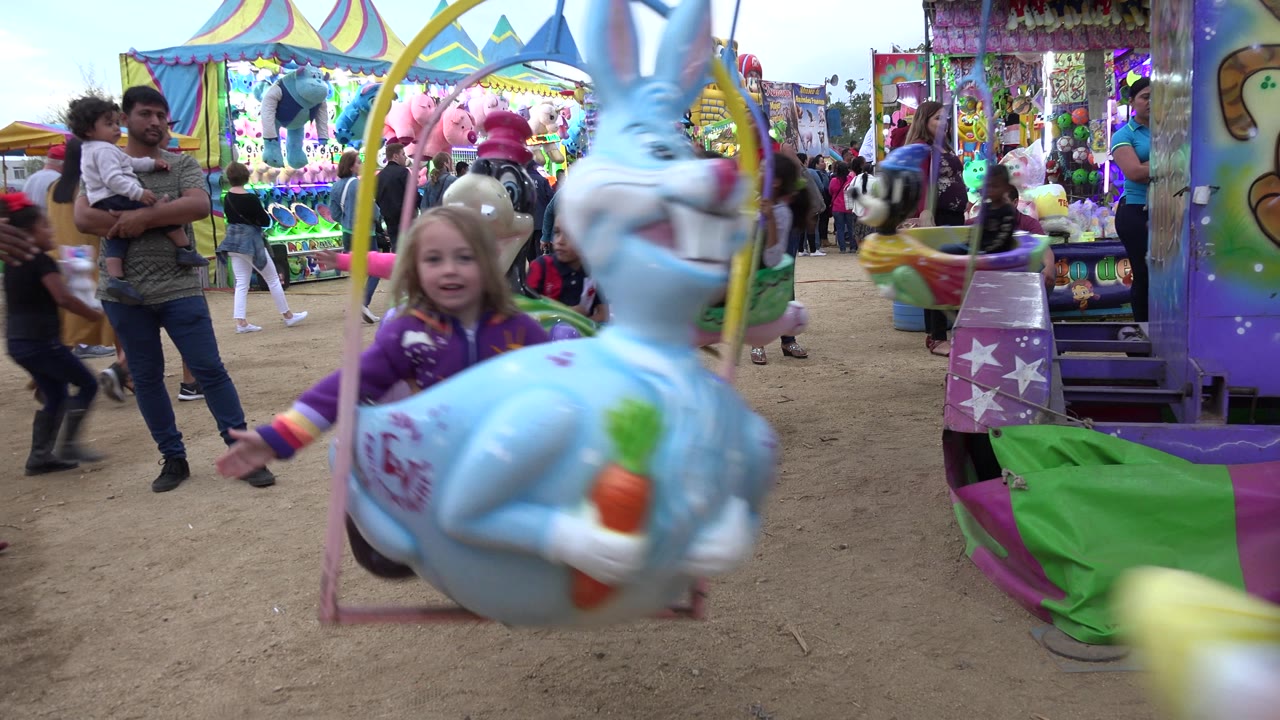 Girl on a blue rabbit carnival seat near adults and bright ride stalls.