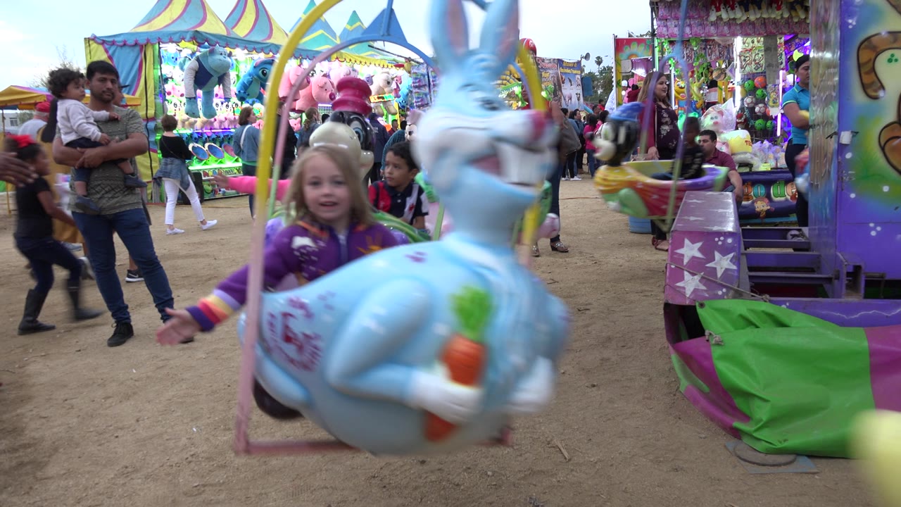The blue rabbit-shaped seat passes close to the lens in San Jose del Cabo, Mexico, while an adult holding a child and carnival booths remain visible in the background.