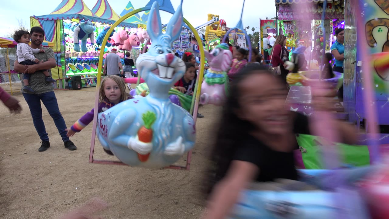 At 0:00 in San Jose Del Cabo, Mexico, a blurred girl rides past the camera while a blue rabbit carousel swing, adults, and stuffed prize booths fill the frame.