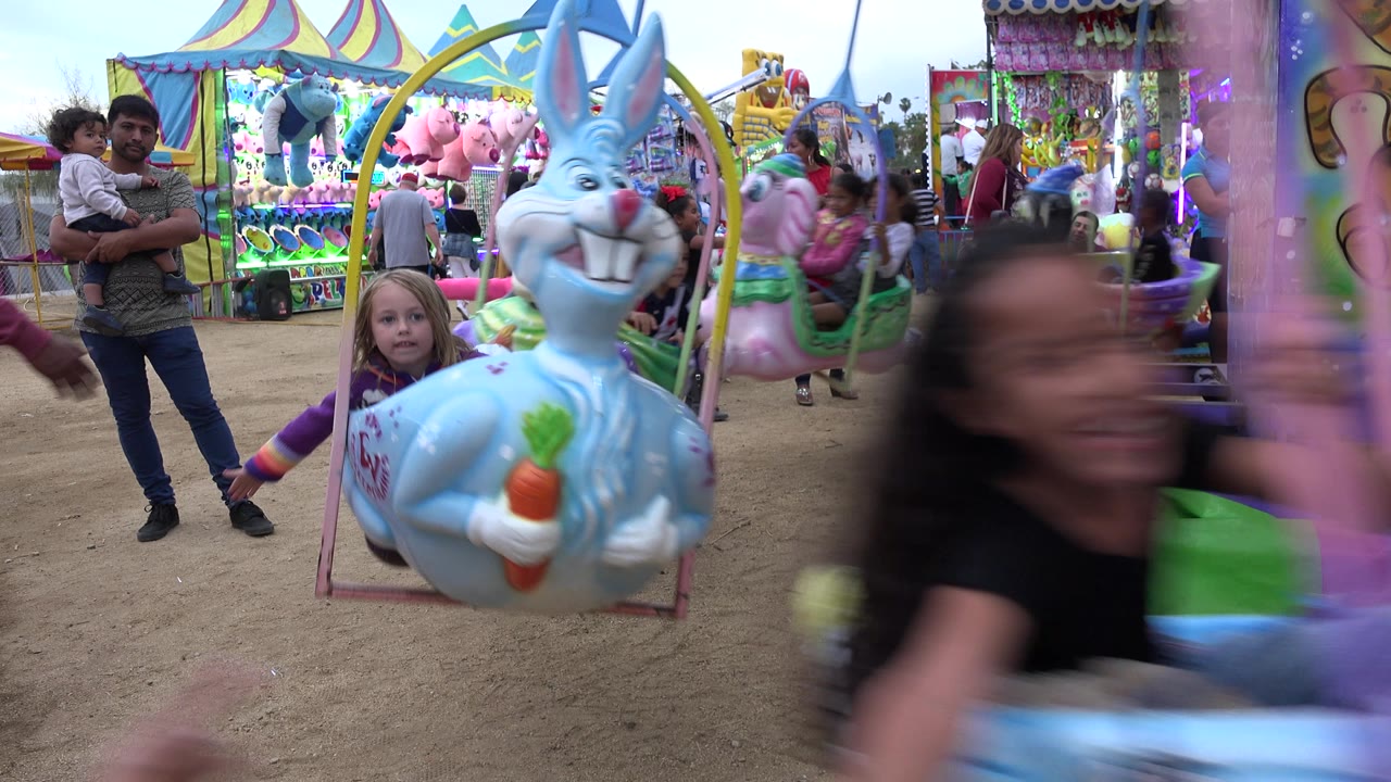 At 0:01 in San Jose Del Cabo, Mexico, the blue rabbit swing is centered while a child blurs through the foreground and adults stand near colorful fair booths.