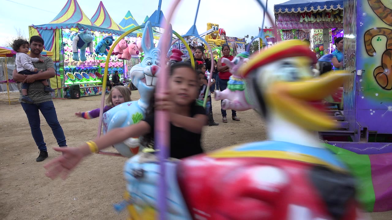 A large duck-shaped carousel seat blurs through the foreground as a child rides behind it in San Jose Del Cabo, Mexico, with stuffed prizes and spectators in the background.