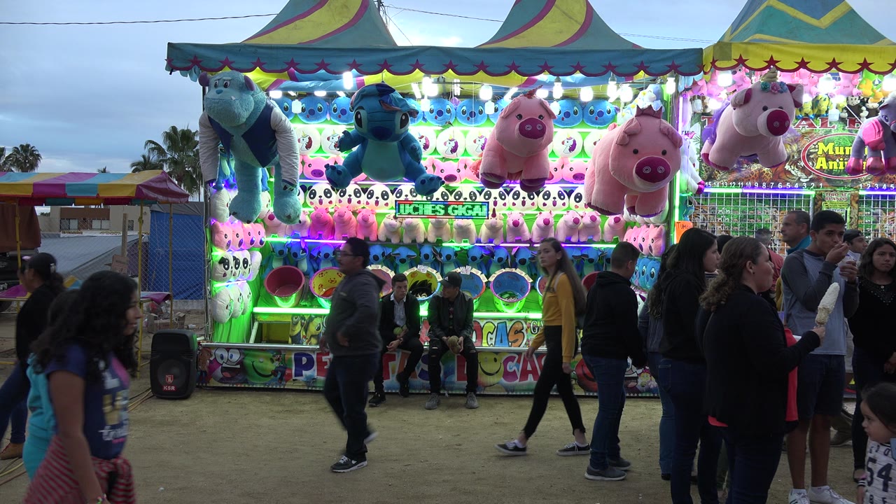 A group of adults and children moves across the foreground while blue and pink plush prizes hang over a lit carnival game booth in San Jose Del Cabo, Mexico.