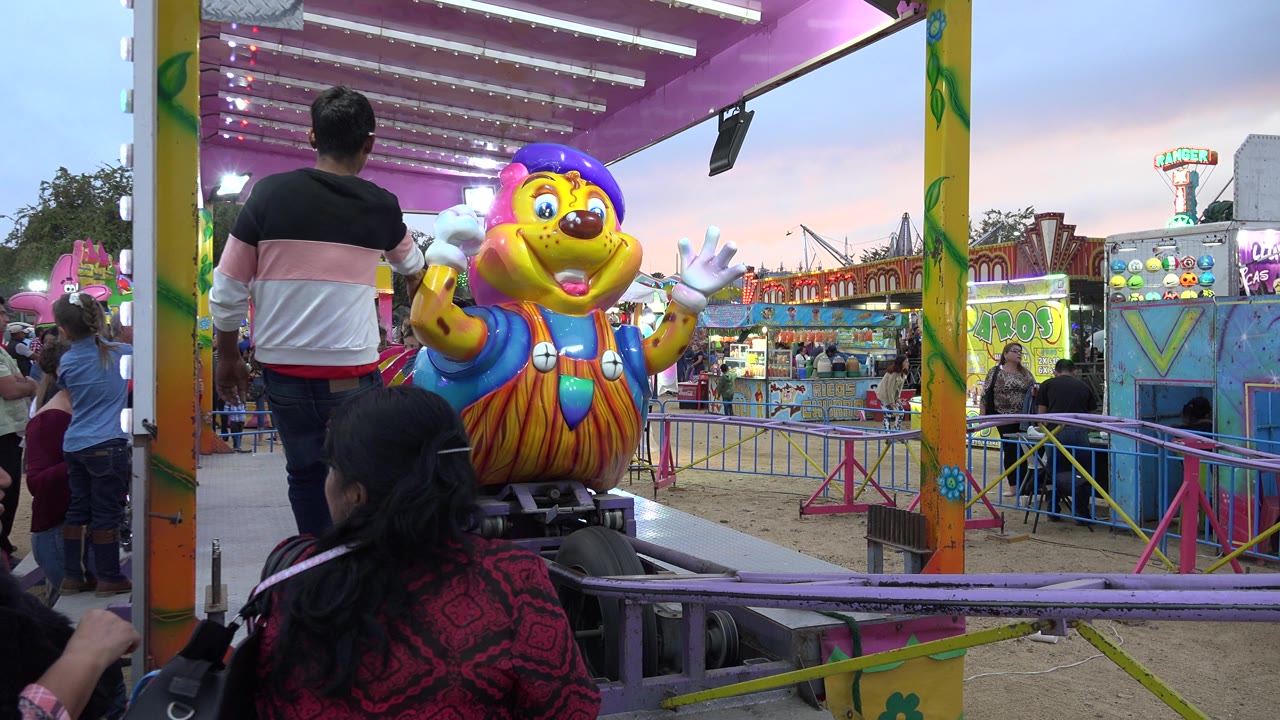 The San Jose del Cabo ride car remains near the platform as an adult stands with his back to camera and fair booths glow in the background.