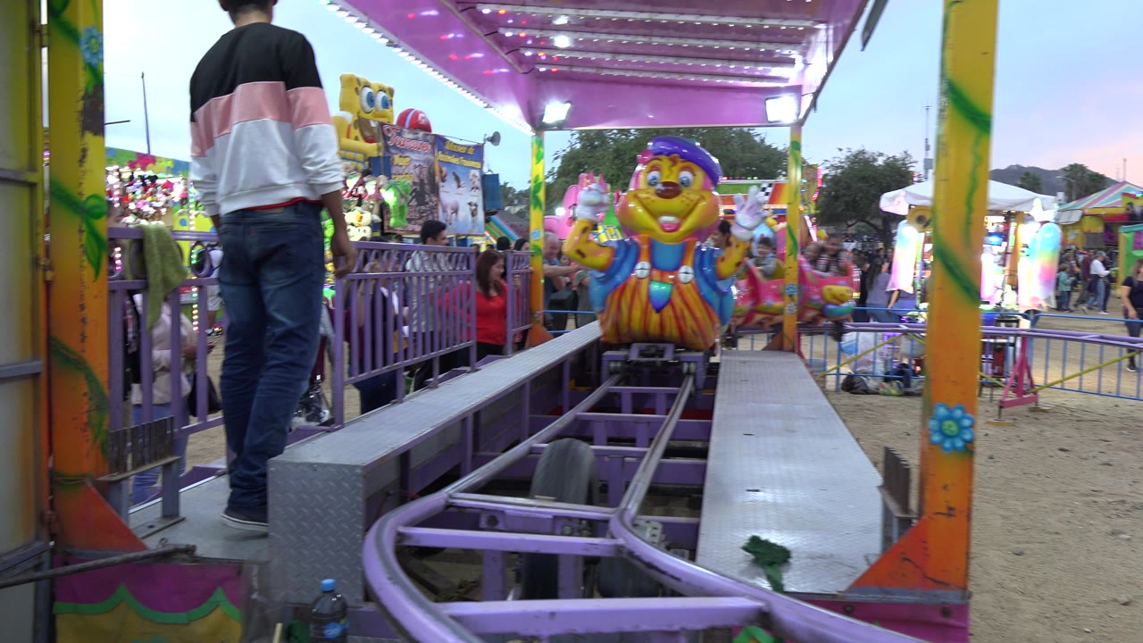 A caterpillar-themed coaster car approaches along purple track at a San Jose del Cabo fair while a man stands beside the loading platform and visitors fill the background.