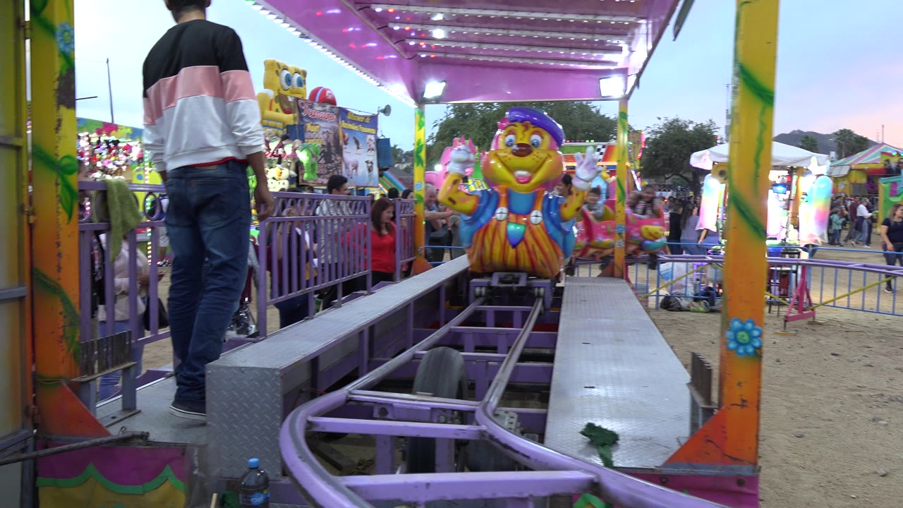 The yellow caterpillar car moves closer on the purple rails under a pink-lit canopy, with a standing adult at left and fairgoers visible beyond the barriers in San Jose del Cabo.