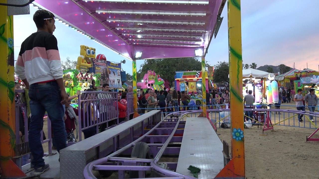 Kiddie roller coaster track beside a carnival platform with people waiting nearby.