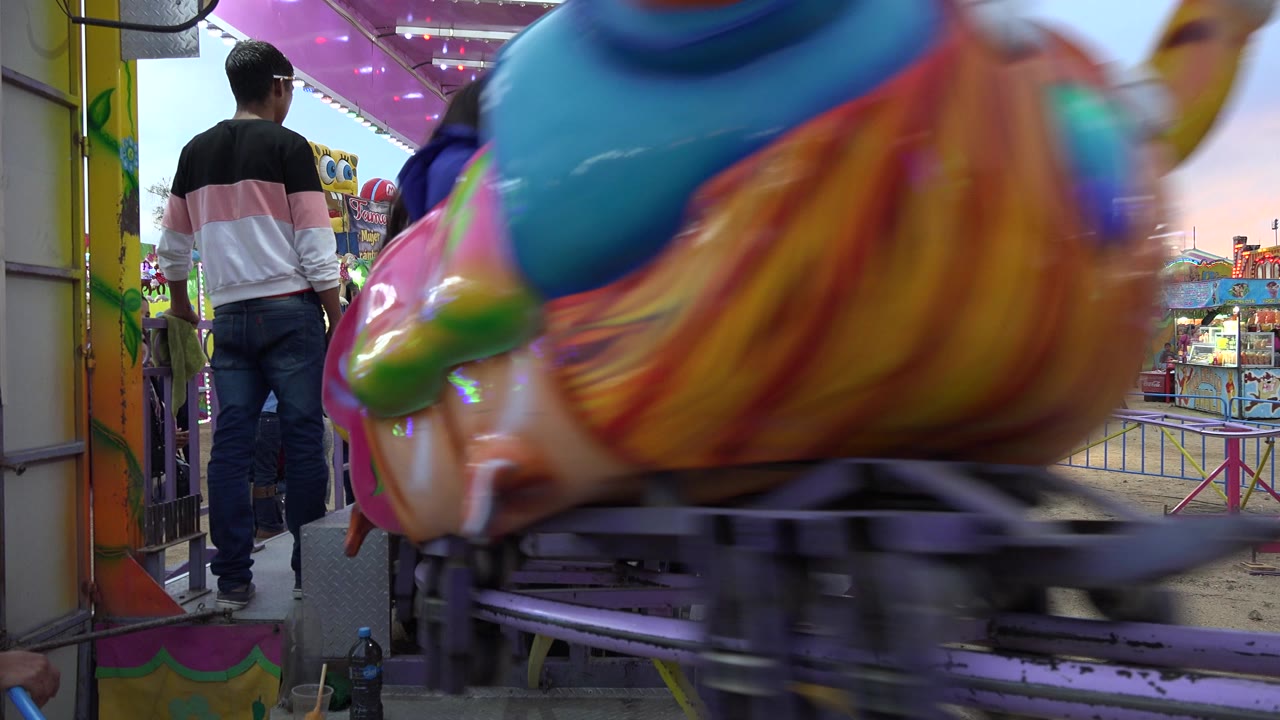 In San Jose del Cabo, a carnival worker stands on the ride platform while a bright animal-shaped car blurs across the foreground and people gather beyond the railings.