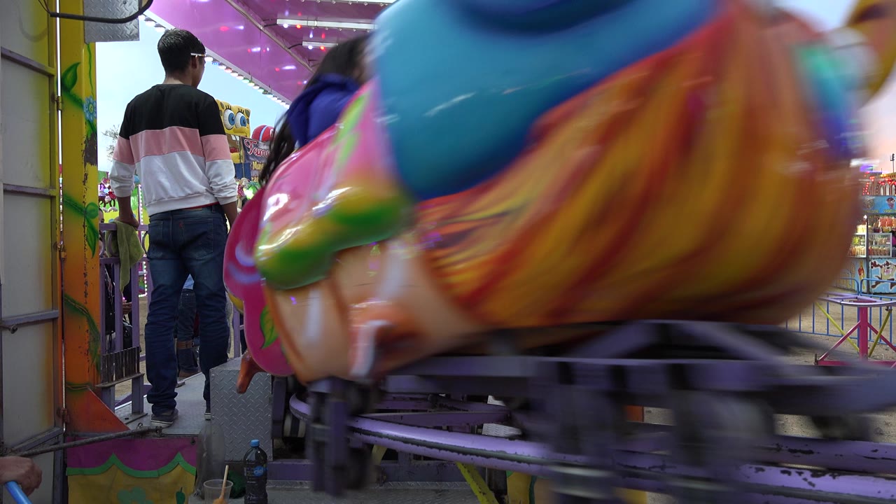 The San Jose del Cabo carnival worker remains near the purple track while a large painted ride car fills the right foreground and riders and visitors are partly visible behind it.