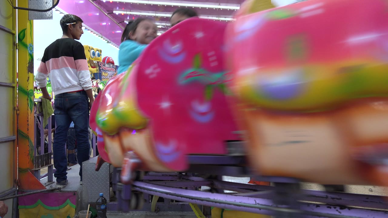 Pink carnival ride car passes in front of a worker and waiting crowd.