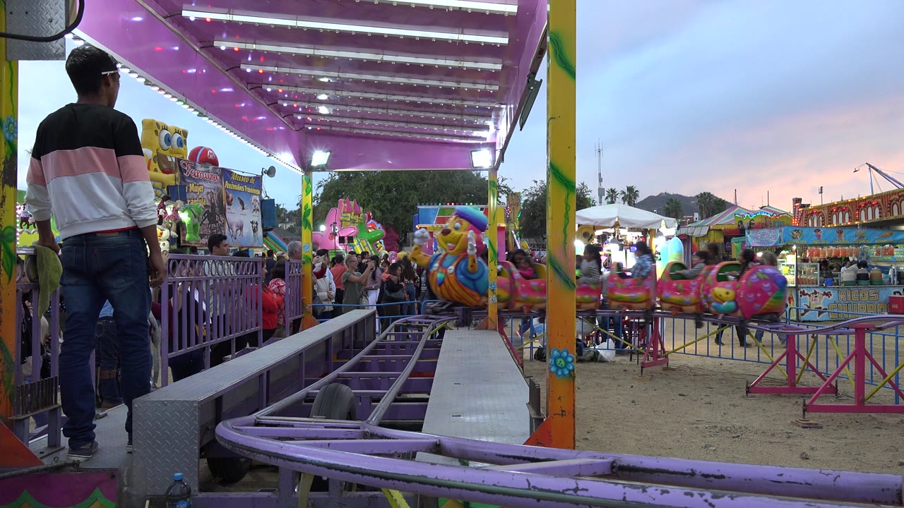 At 0:01 in San Jose del Cabo, the view opens across the ride enclosure toward bright fair stalls, railings, and small character cars under a blue and pink evening sky.
