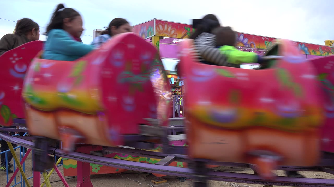 A pink amusement ride car passes across the foreground in San Jose Del Cabo, Mexico, partially obscuring people and bright prize booths behind it.